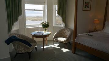 A bedroom with a four-poster bed, a chair, a small table with a vase of flowers, and a window with a view of the sea.