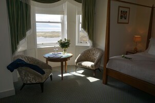Un dormitorio con cama con dosel, una silla, una mesita con un florero y un ventanal con vistas al mar.