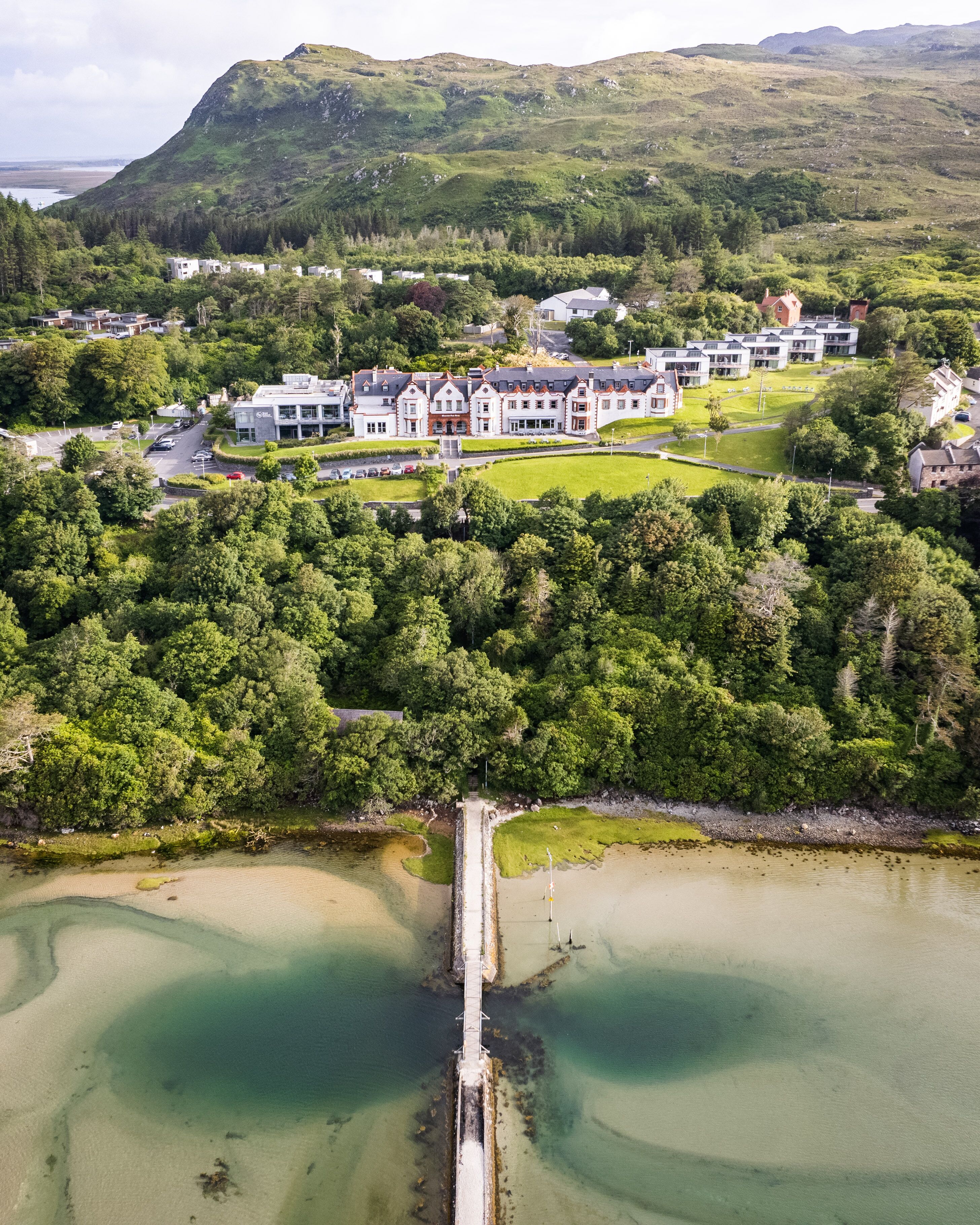 An aerial view of a coastal area with a large building complex surrounded by dense greenery and a pier extending into a calm bay.