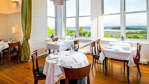 A dining area with tables set for a meal, large windows offering a view of the sea and greenery, and pendant lights hanging above the tables. - Mulranny Park Hotel (Mulranny)