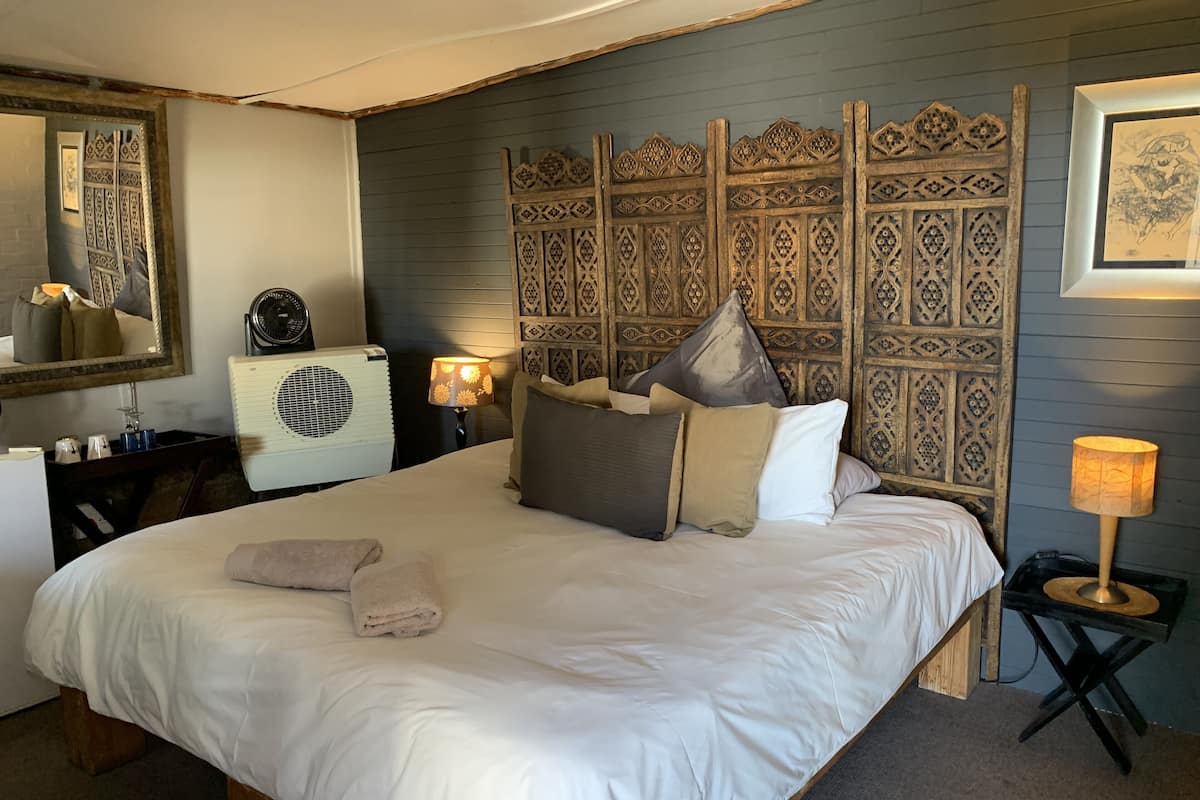 Una cama bien hecha con sábanas blancas y varias almohadas, una mesita de noche con lámpara, un cabecero decorativo y un espejo en la pared.