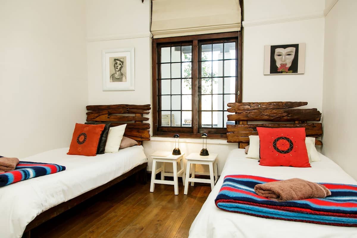 Una habitación con dos camas, cabeceros de madera, ropa de cama blanca, una ventana con persianas y una pequeña mesa con lámparas.