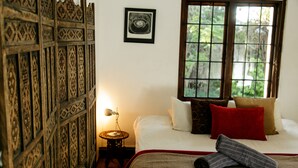 Een slaapkamer met een bed, kussens, een deken, een nachtkastje, een lamp en een raam met uitzicht op groen.