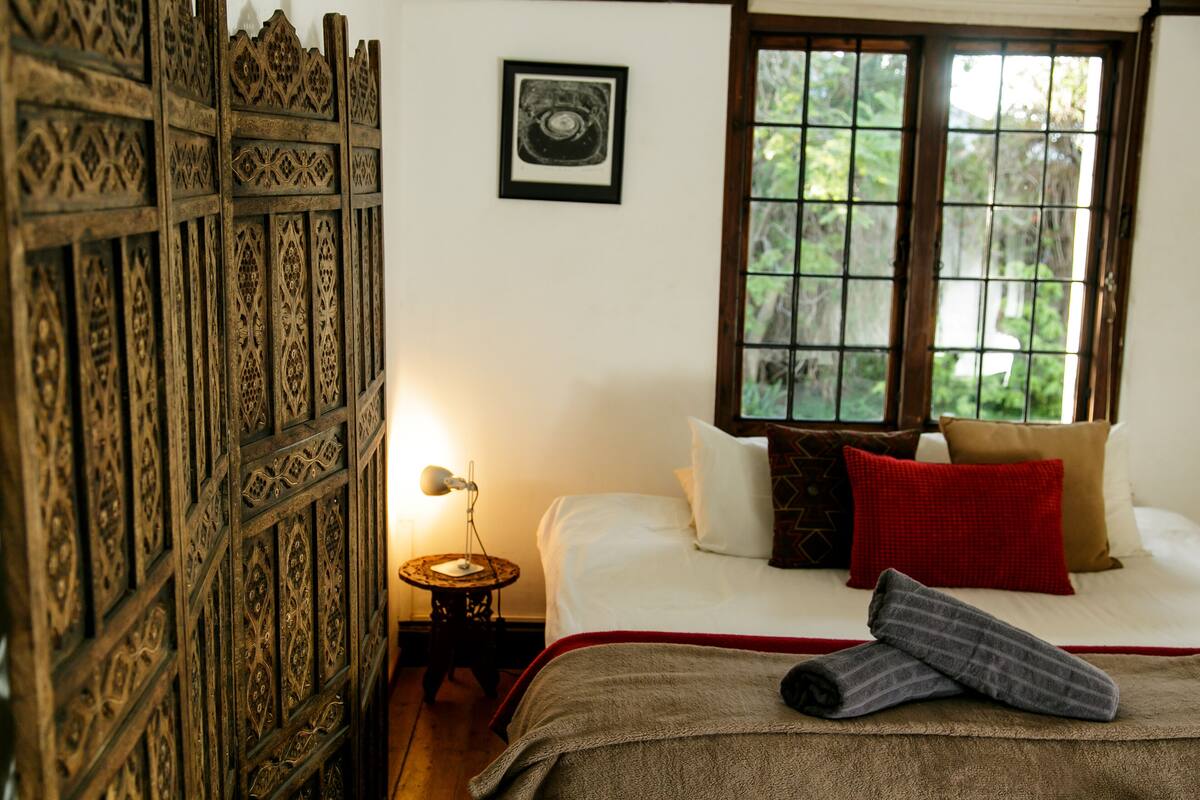 Un dormitorio con cama, almohadas, una colcha, una mesita de noche, una lámpara y una ventana con vista a la vegetación.