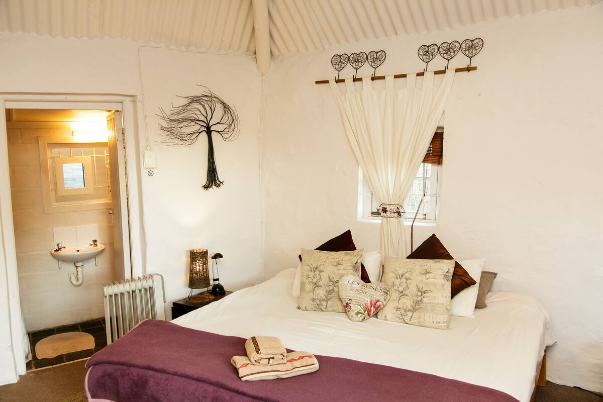 Un dormitorio con cama, almohadas, ventana con cortinas, radiador, mesita de noche, lámpara y una obra de arte en la pared.