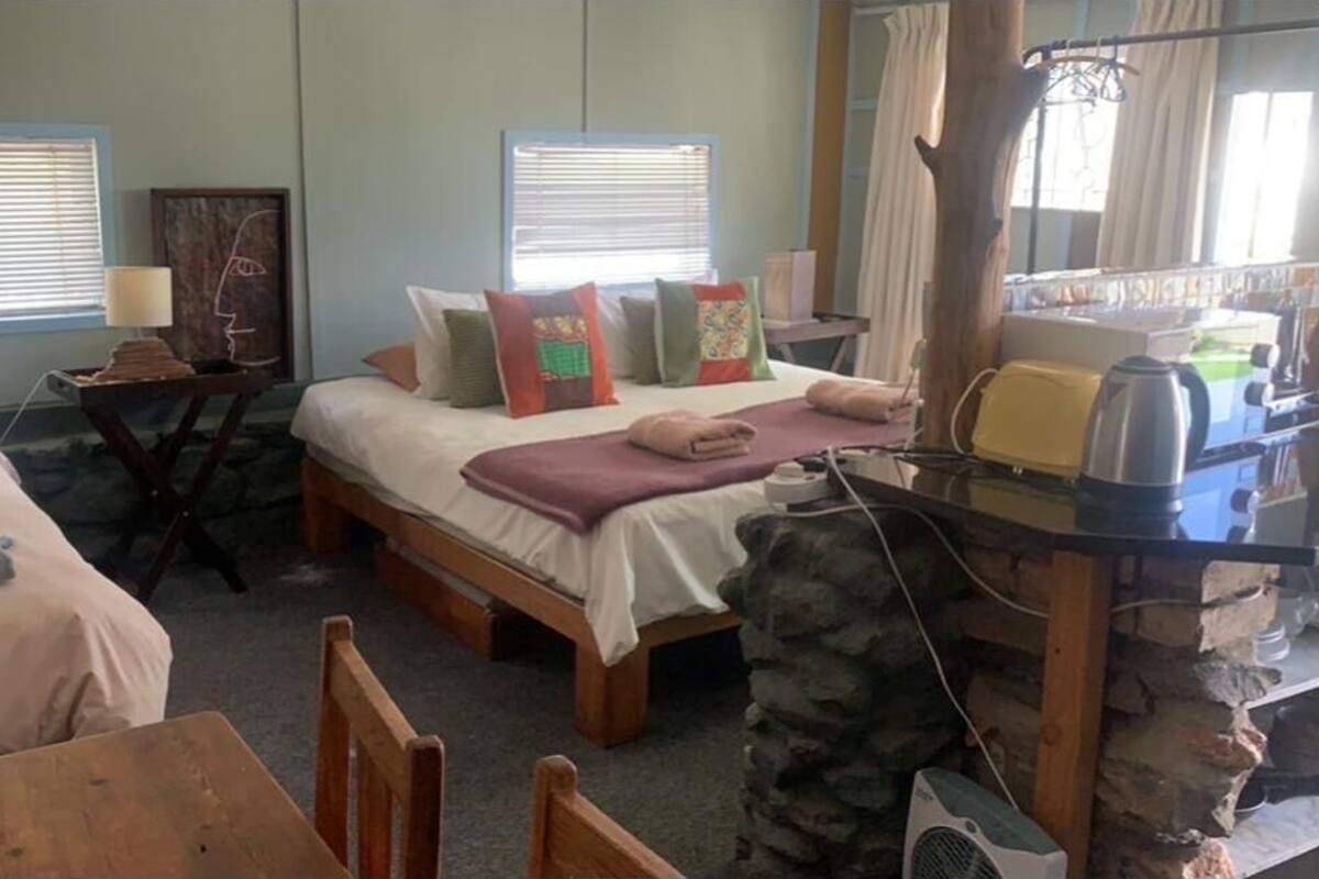 Habitación con dos camas, un escritorio y una silla. Cuenta con ventanas con persianas, una lámpara y un cuadro en la pared.