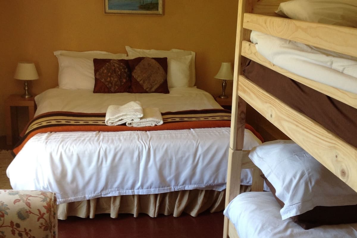 A hotel room with a bunk bed, a single bed, a chair, and a painting on the wall.