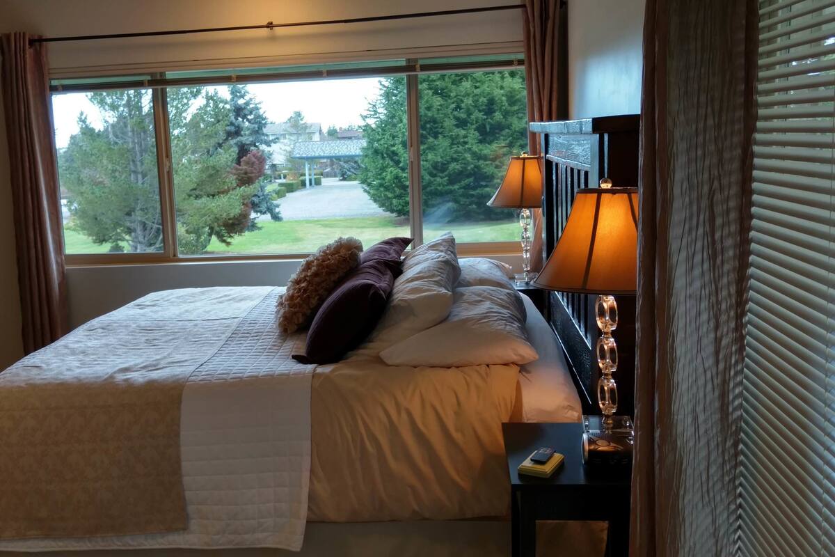 A bedroom with a large bed, a bedside table with a lamp, and a view of a green landscape through a window.