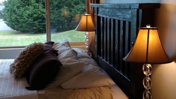 Una cama bien tendida con ropa de cama blanca y beige, una almohada decorativa oscura y una almohada decorativa. Dos lámparas de pared con luz cálida. Una ventana muestra vistas de un césped verde y árboles.