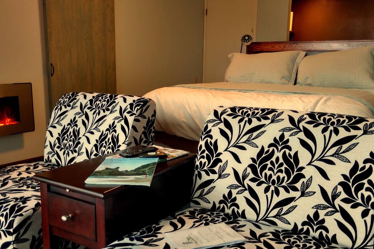 A hotel room with a bed, two upholstered chairs with a floral pattern, a bedside table with a book and a remote control, and a wall-mounted fireplace.