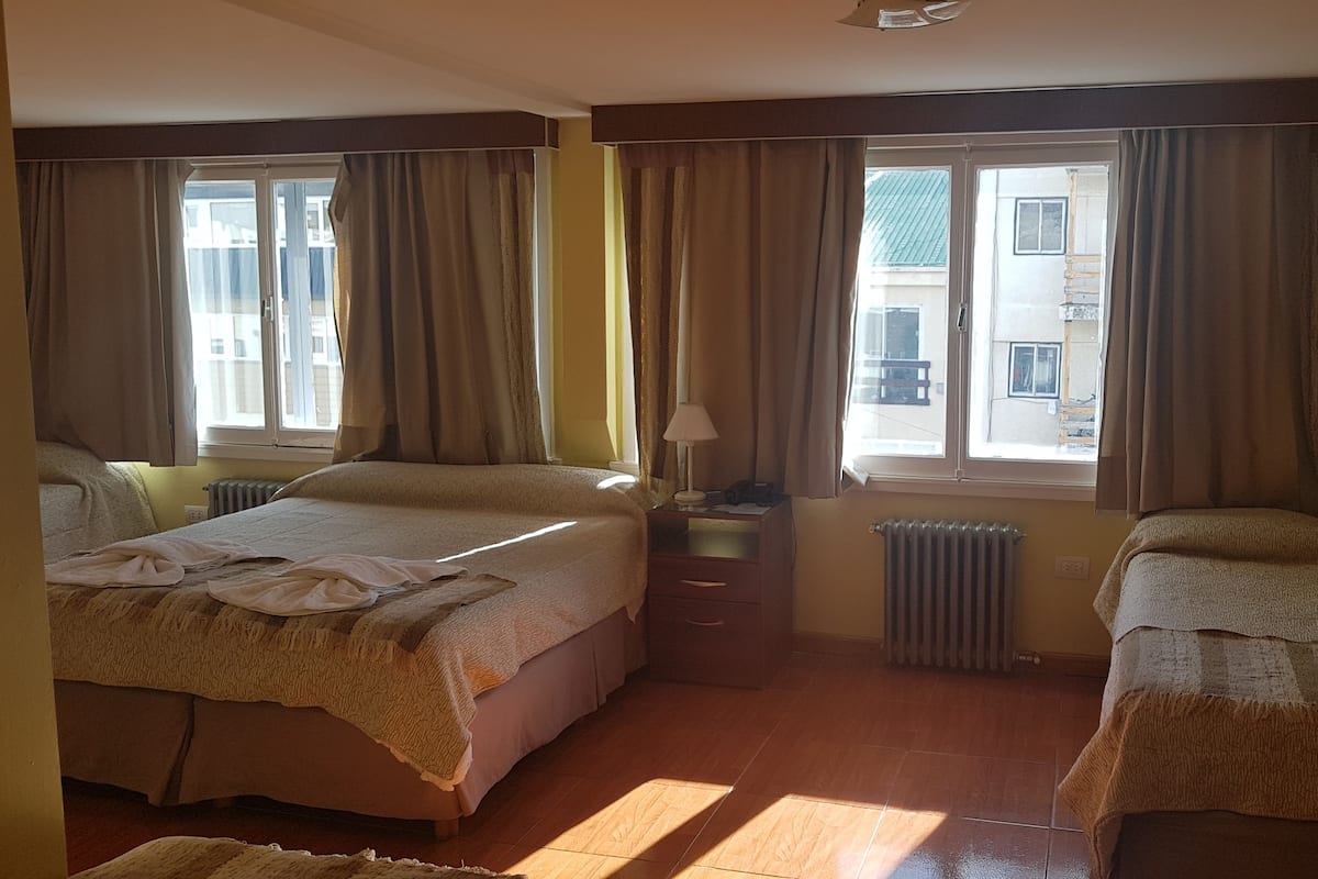 A hotel room with two beds, a nightstand, a lamp, and a view of the outside.