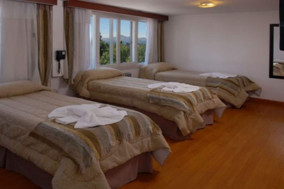 Camera d'albergo con tre letti singoli, ognuno con un piumone e un asciugamano bianco sopra. La stanza ha il pavimento in legno e una finestra con tende.