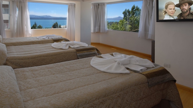 A hotel room with three beds, large windows with curtains, and a view of a lake and mountains. - Hotel Monte Cervino (San Carlos de Bariloche)