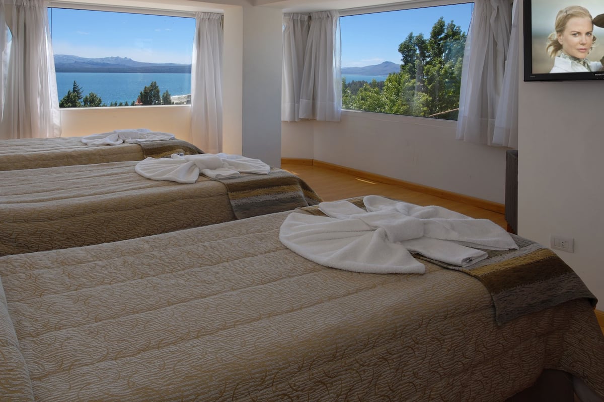 Una camera d'albergo con tre letti, ampie finestre con tende e vista su un lago e montagne.