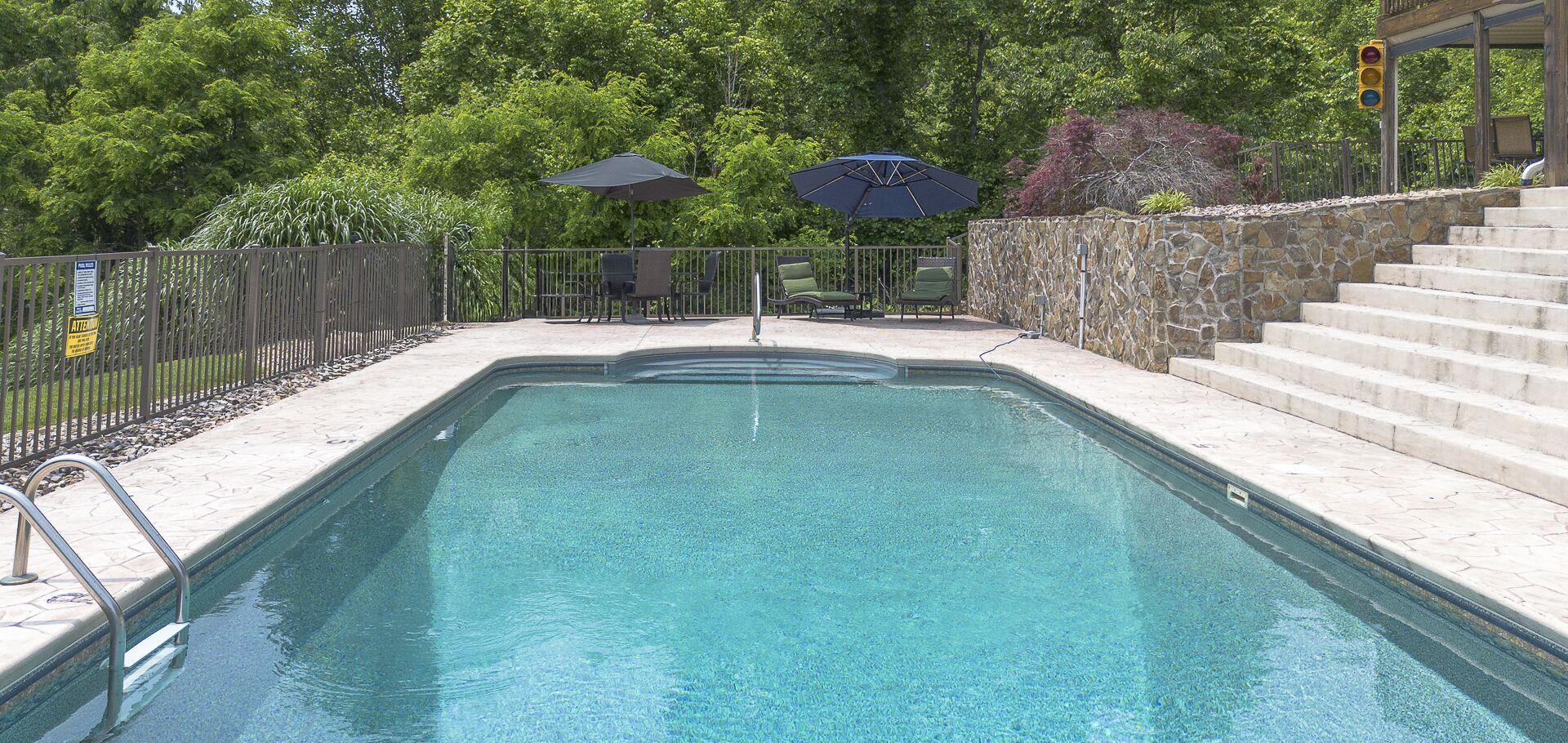 A rectangular swimming pool with a stone wall and steps leading up to a covered area with umbrellas.
