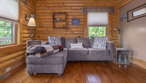 A cozy cabin interior with a sectional sofa, a lamp, a small table, and a window with blinds. - Shirley's Bed and Breakfast (Roanoke)