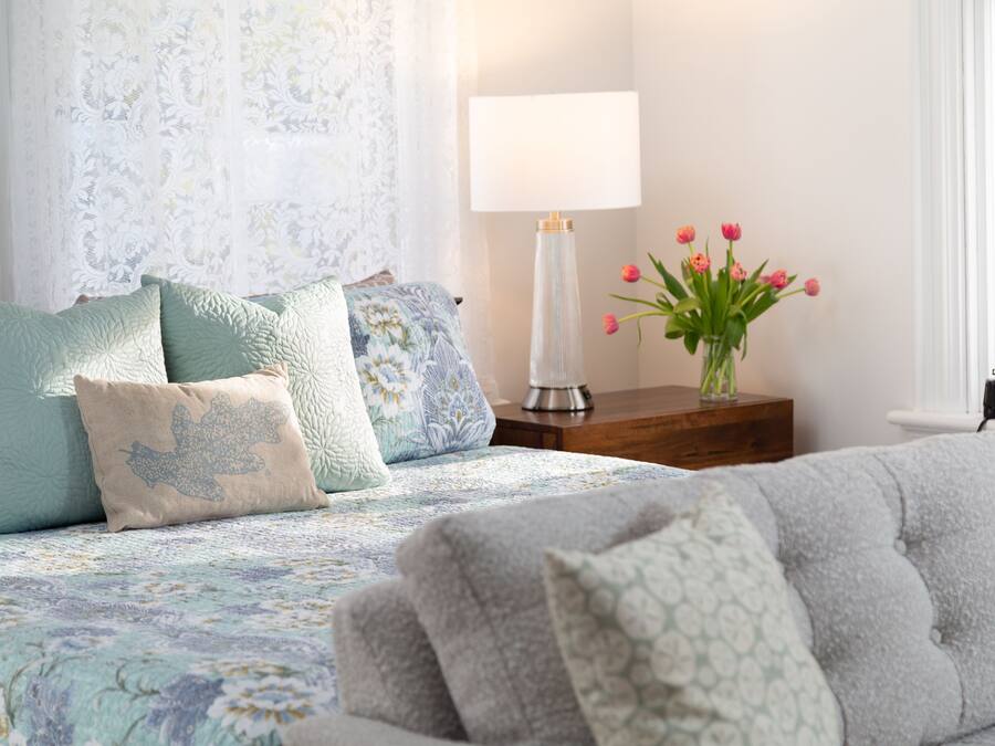 Quarto com colcha florida, várias almofadas, abajur e um vaso com flores.