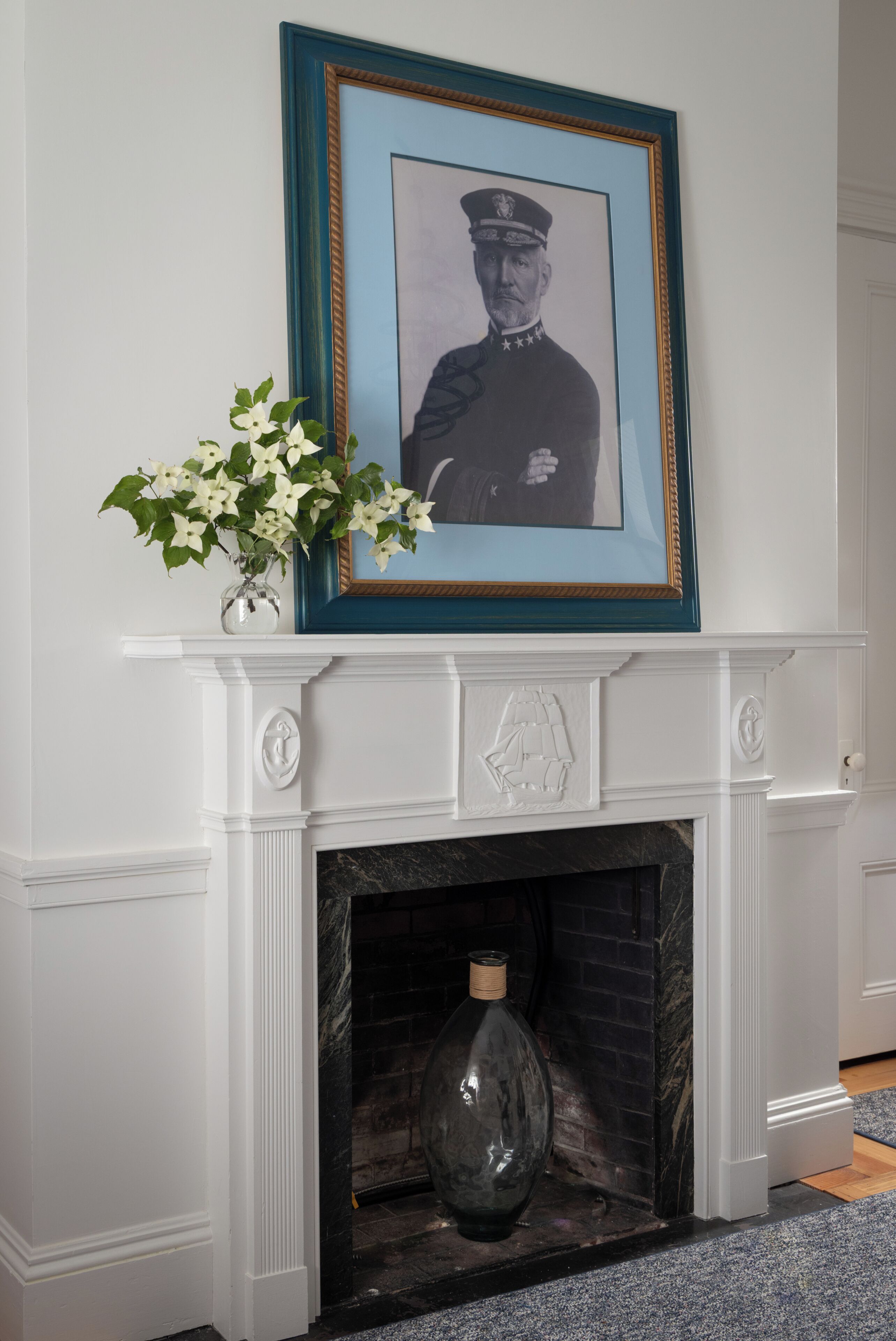 A fireplace with a framed portrait above it, a vase with flowers to the left, and a bottle in the fireplace.