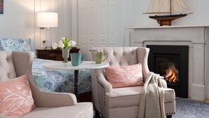Une pièce chaleureuse avec une cheminée, deux fauteuils, une petite table ornée d’un vase de fleurs et une décoration représentant un voilier au-dessus de la cheminée.