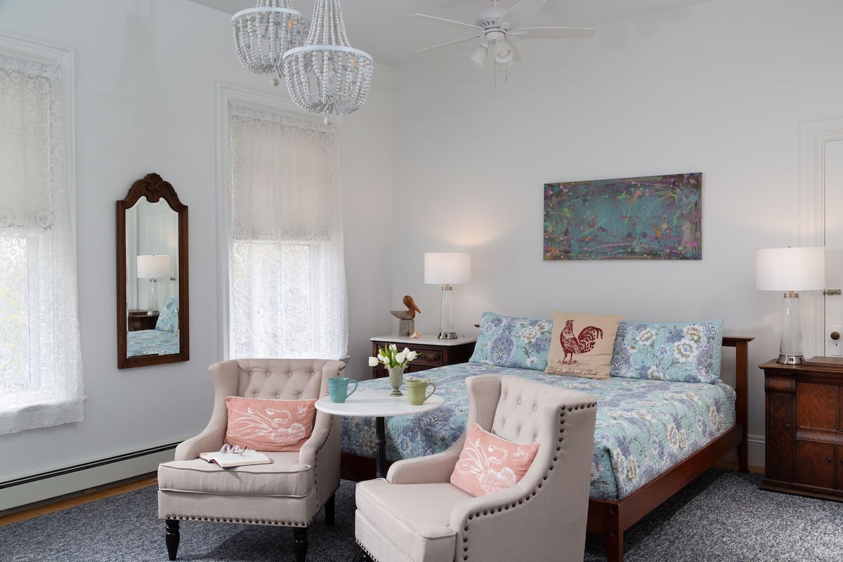 Une chambre à coucher comprenant un lit, deux fauteuils, une petite table, un lustre, un ventilateur de plafond et un tableau accroché au mur.