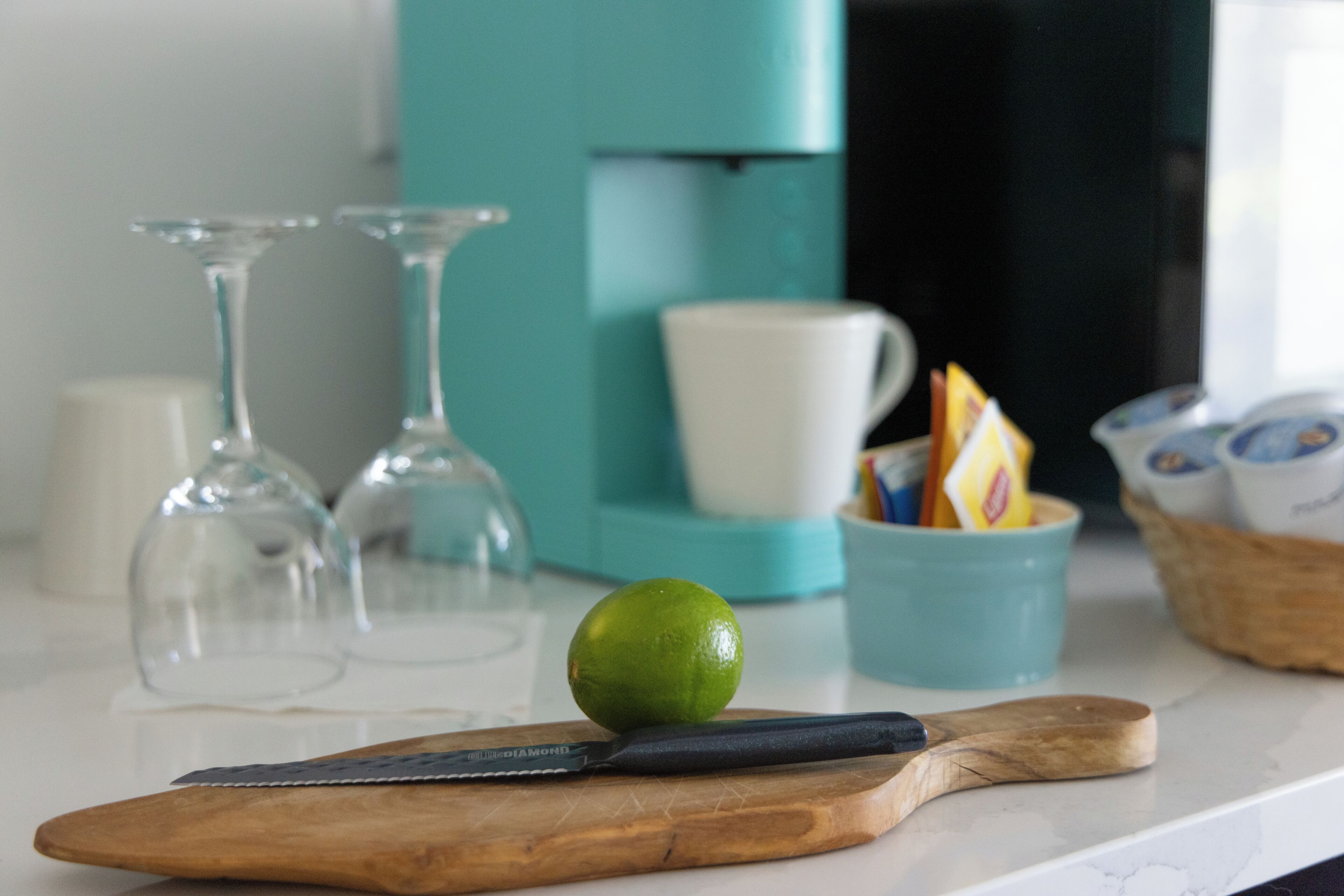 Uma bancada de cozinha com um limão, uma faca, uma tábua de corte de madeira, um eletrodoméstico azul-turquesa e uma caneca branca.