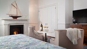 Une chambre avec une cheminée, un lit recouvert d’une courtepointe à motifs floraux, une petite table avec une bouteille et des verres, et un téléviseur fixé au mur.