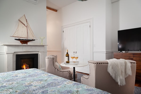 A bedroom with a fireplace, a bed with a floral bedspread, a small table with a bottle and glasses, and a television on the wall.