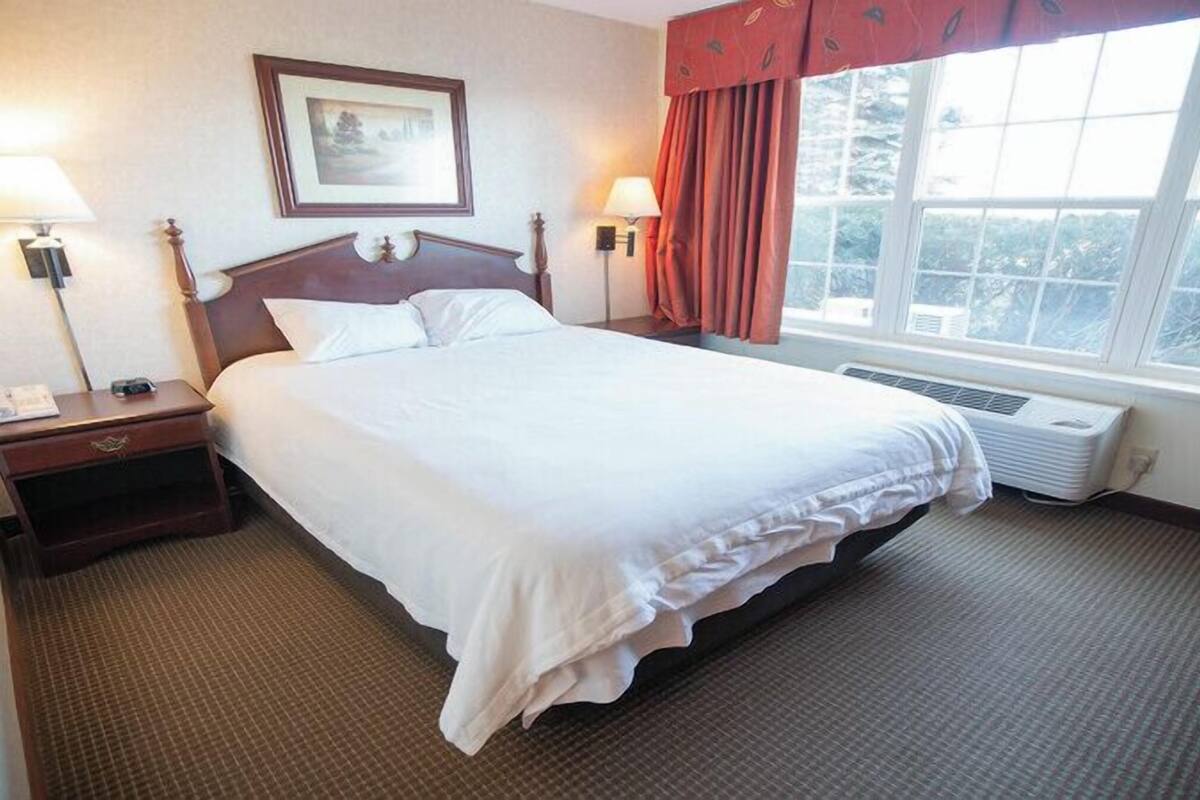 Ein Hotelzimmer mit einem großen Bett, zwei Nachttischlampen, einem gerahmten Bild an der Wand, einem Fenster mit Vorhängen und einem Heizlüfter unter dem Bett.
