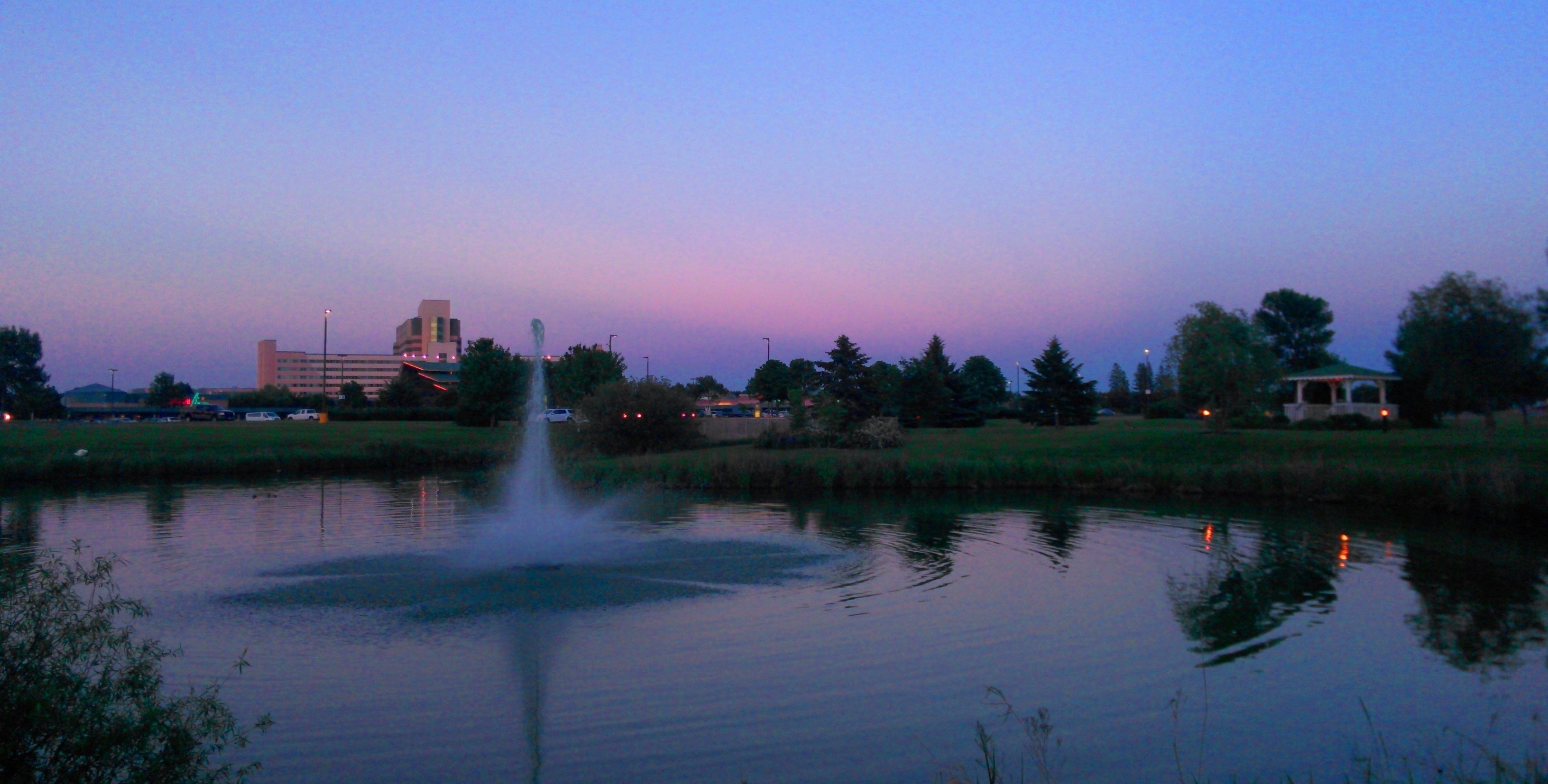A calm lake with a fountain in the center, surrounded by trees and a building in the background.