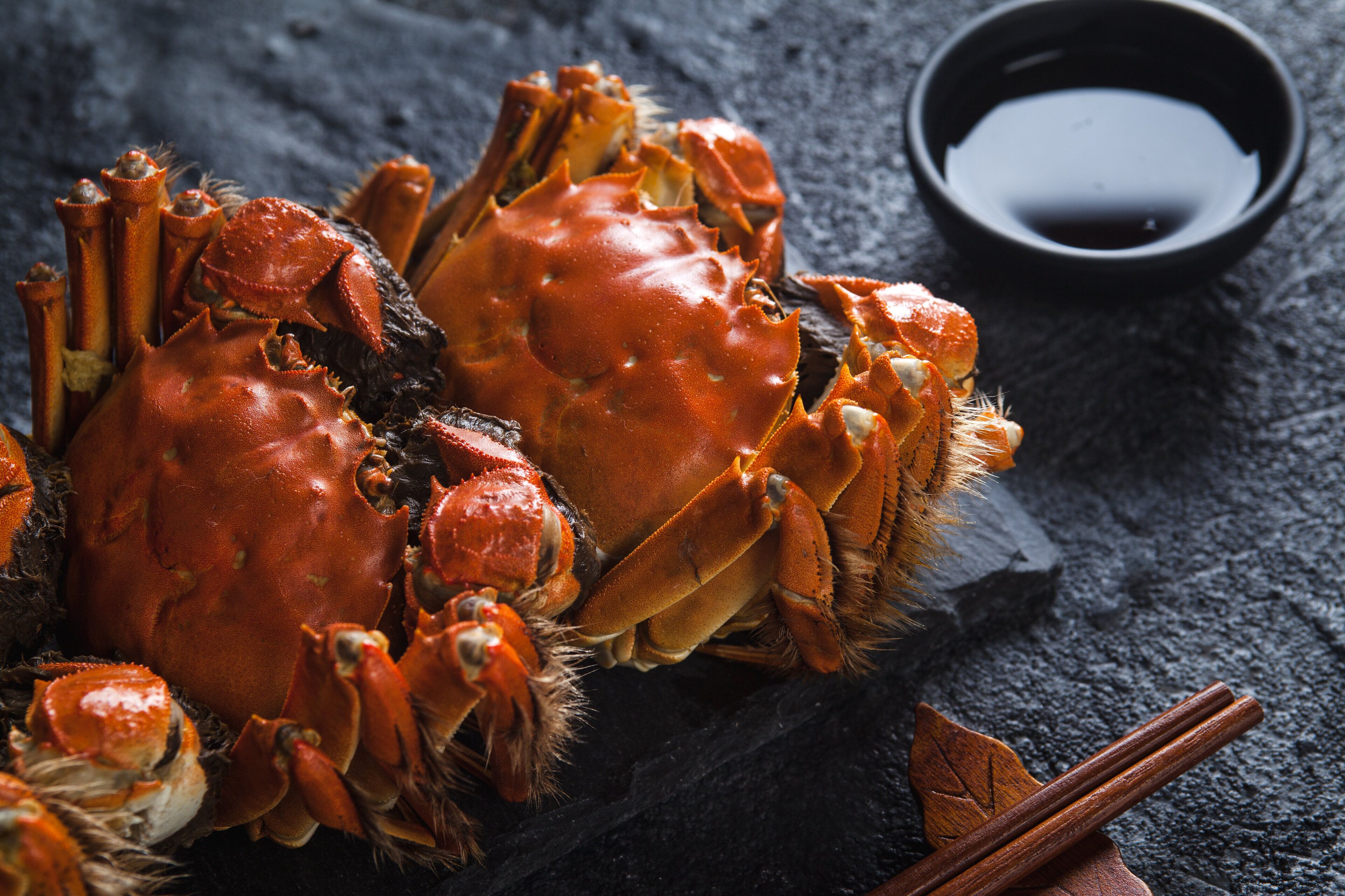 A crab with a bowl of sauce and chopsticks on a dark surface.