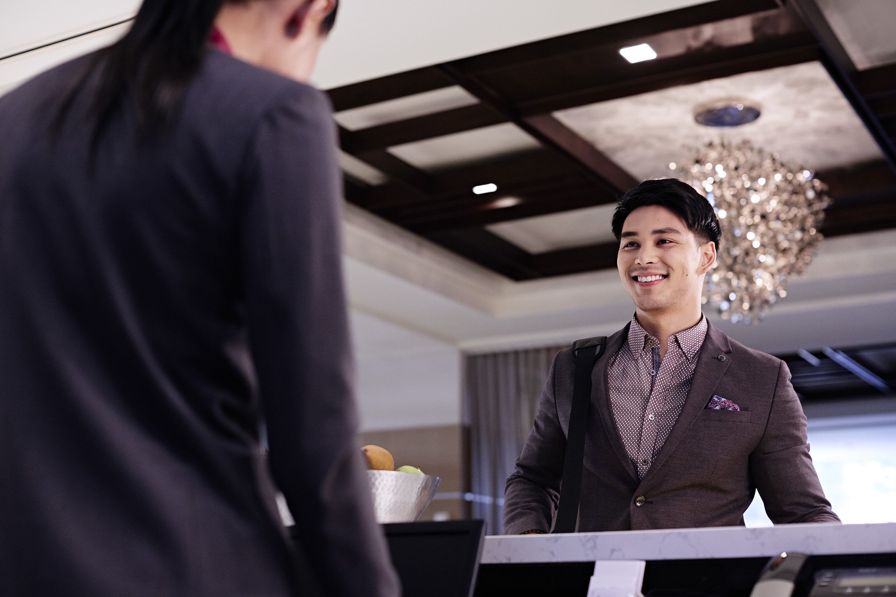 A man in a suit standing at a reception desk in a hotel lobby with a chandelier and a fruit bowl in the background.