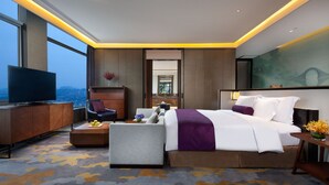Una habitación de hotel moderna con una cama grande, un televisor de pantalla plana, una zona de estar con sofá y sillón, y vistas a la ciudad a través de amplios ventanales.