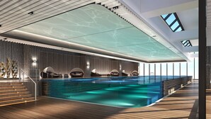 Una piscina interior moderna con techo de cristal, cubierta de madera y sillones de descanso.