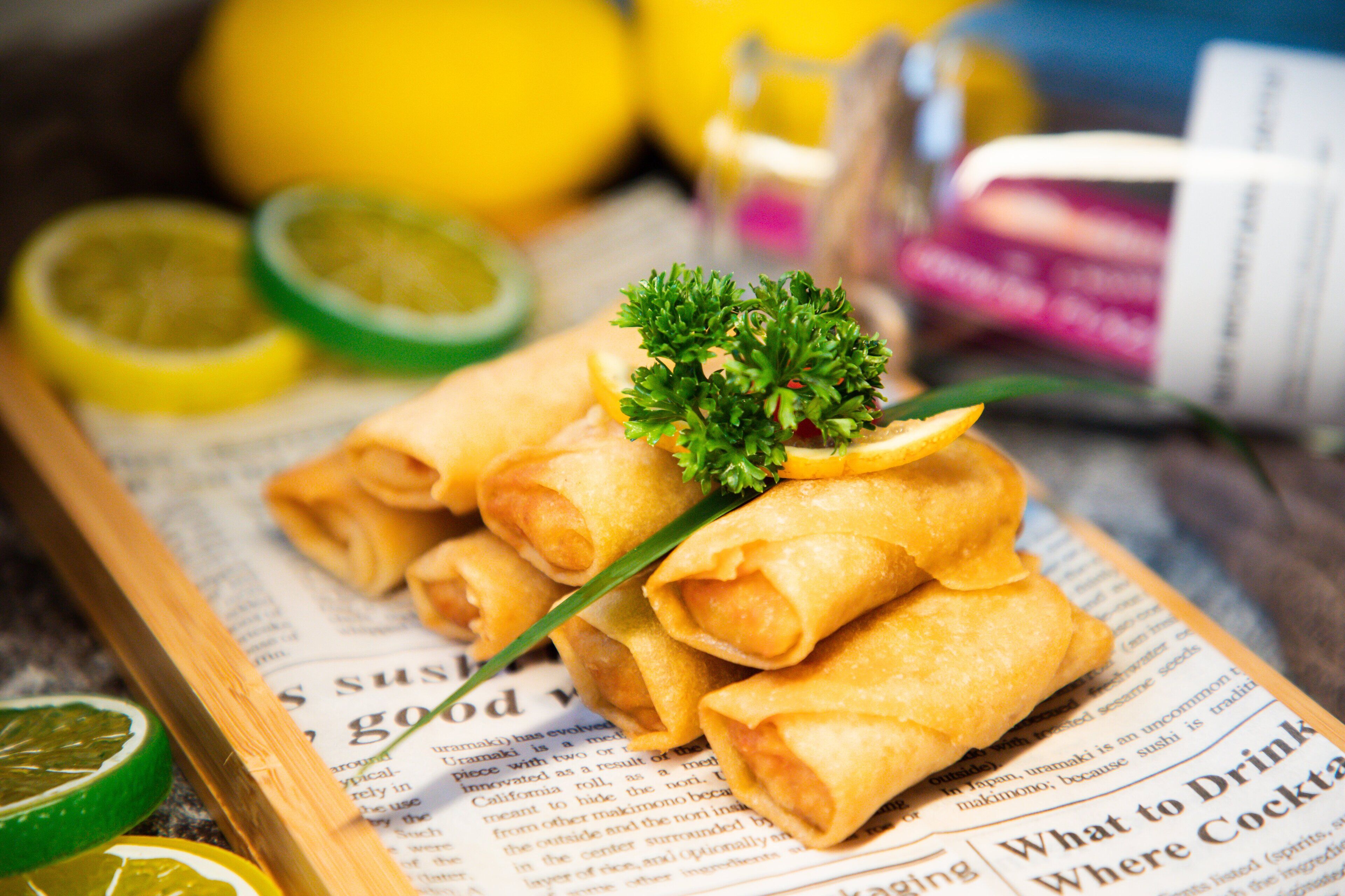 A tray of spring rolls on a newspaper with lemon slices and a sprig of parsley.