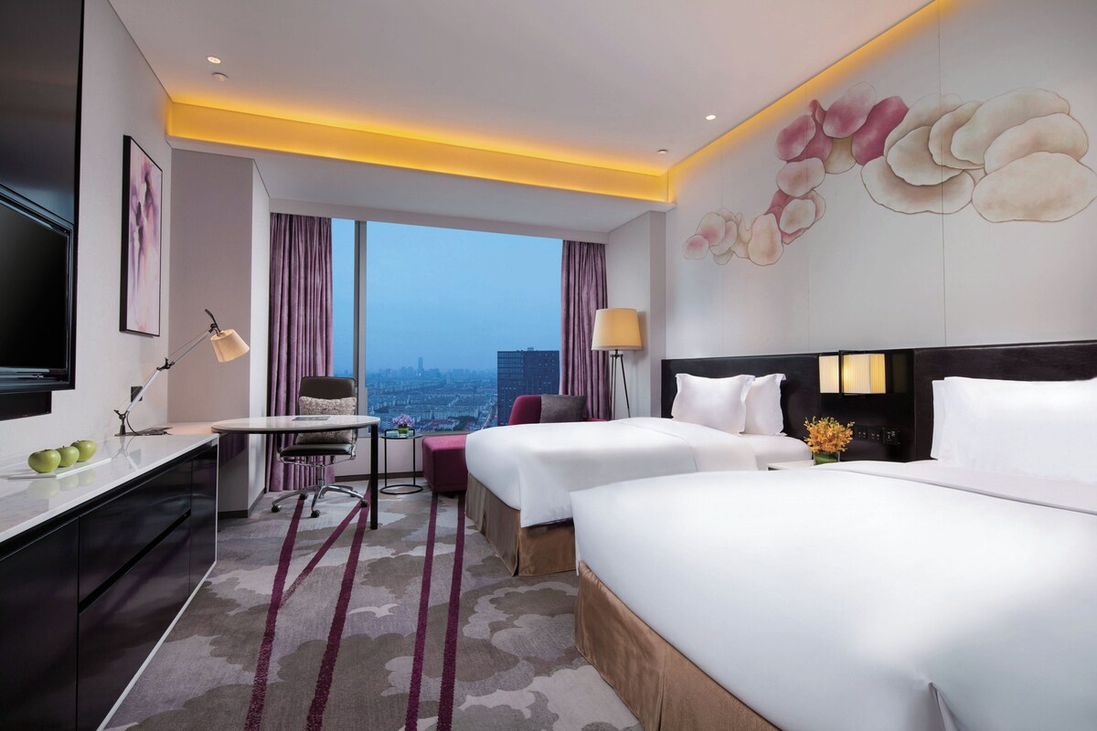 A hotel room with two beds, a desk, a chair, and a view of the city.