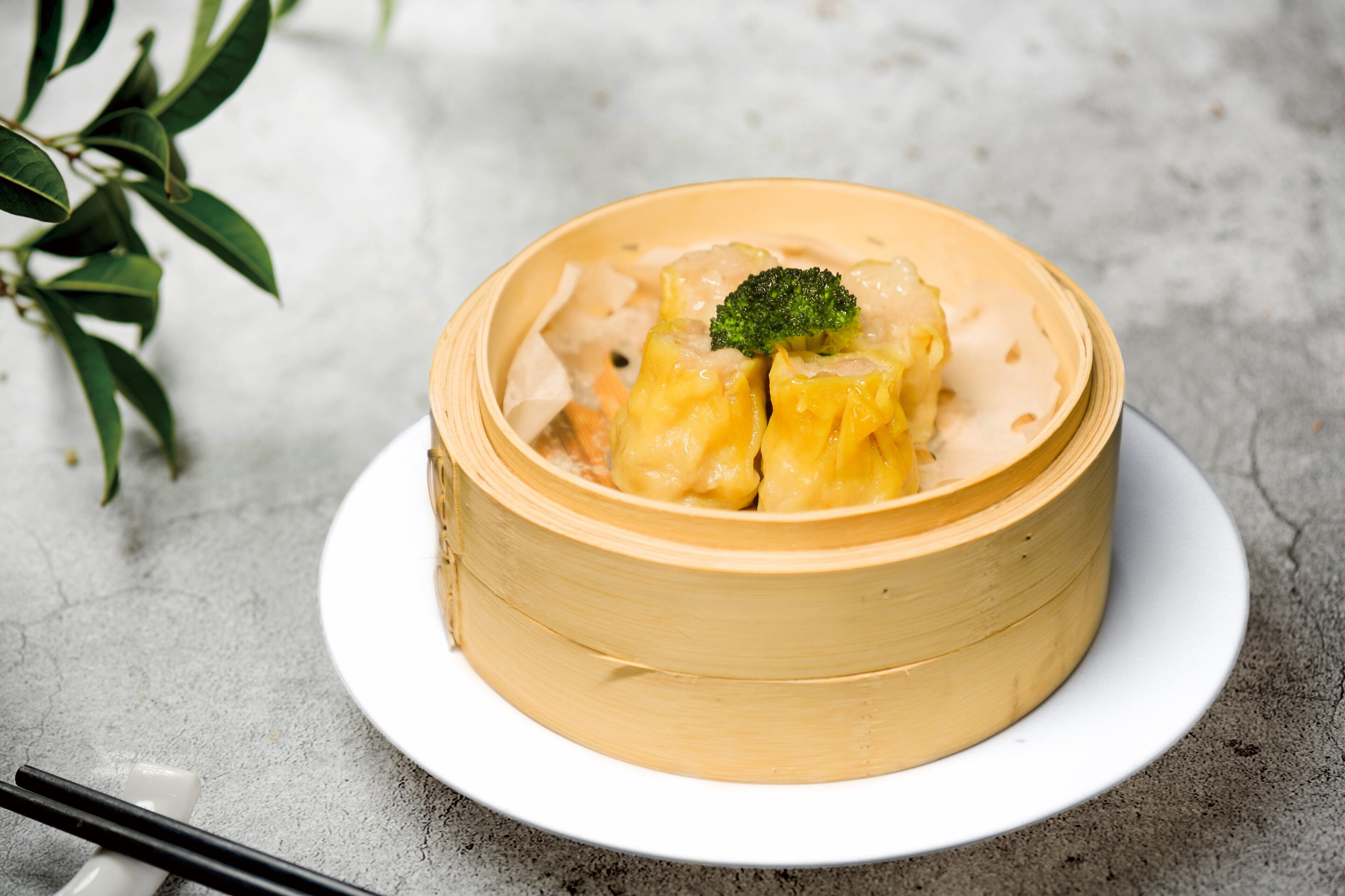 A bamboo steamer with dumplings and a broccoli garnish on a white plate.
