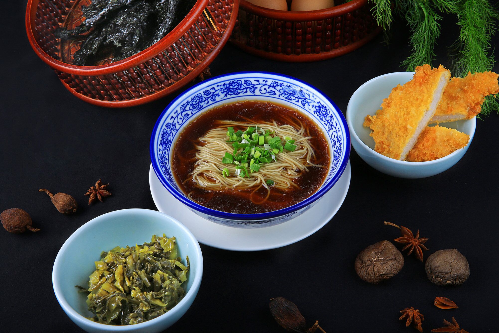 Un tazón de sopa de fideos con cebolletas, rodeado de diversos ingredientes como algas secas, anís estrellado y un tazón de verduras encurtidas.