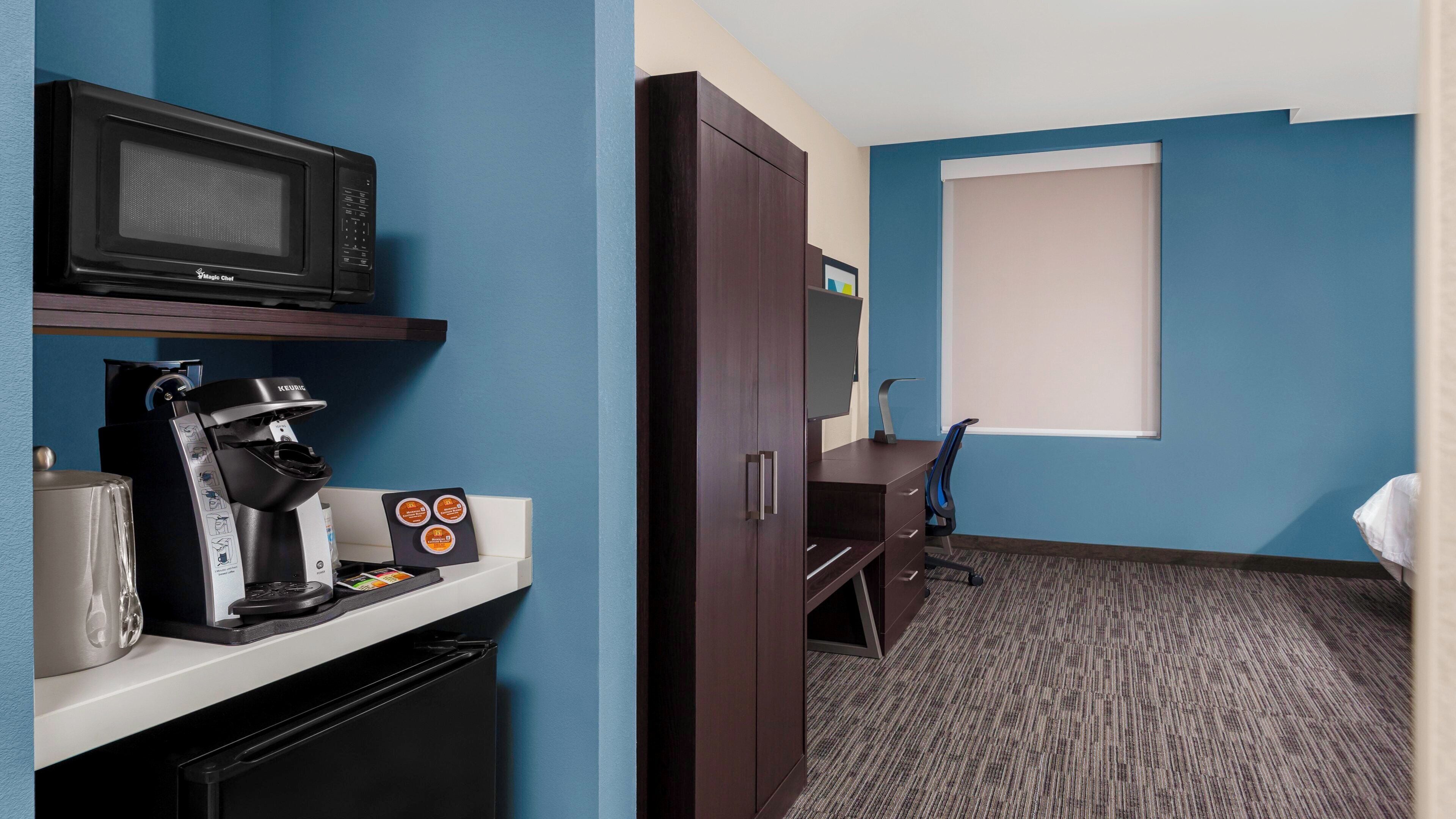 A hotel room with a kitchenette, a microwave, a coffee machine, a wardrobe, a desk, and a large window.
