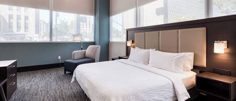 Habitación de hotel con una cama grande, un escritorio, una silla y vistas de la ciudad a través de amplias ventanas.