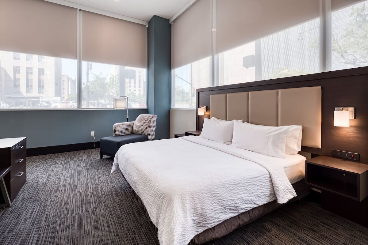 A hotel room with a large bed, a desk, a chair, and a view of the city through large windows.