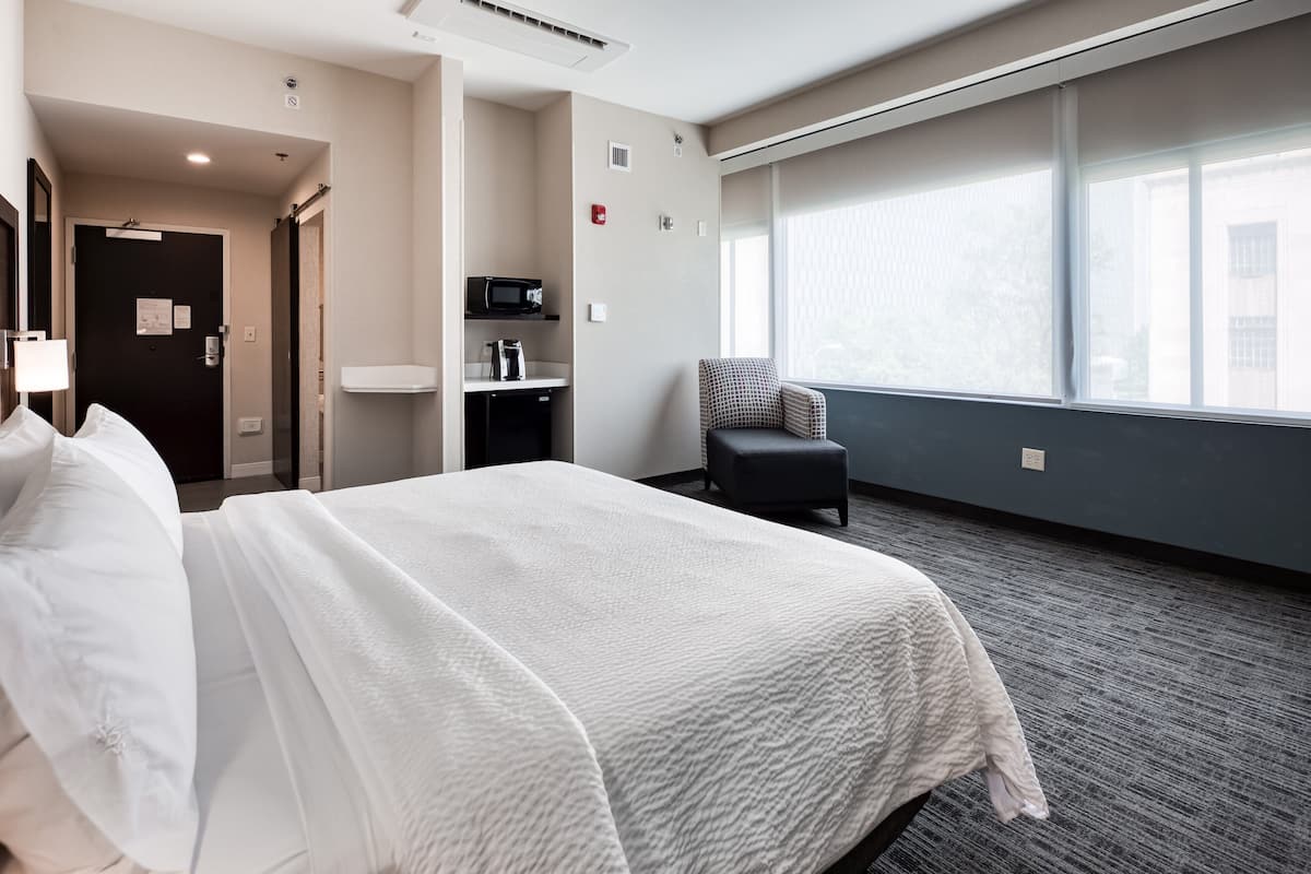 A hotel room with a bed, a chair, a desk with a microwave, and a large window.