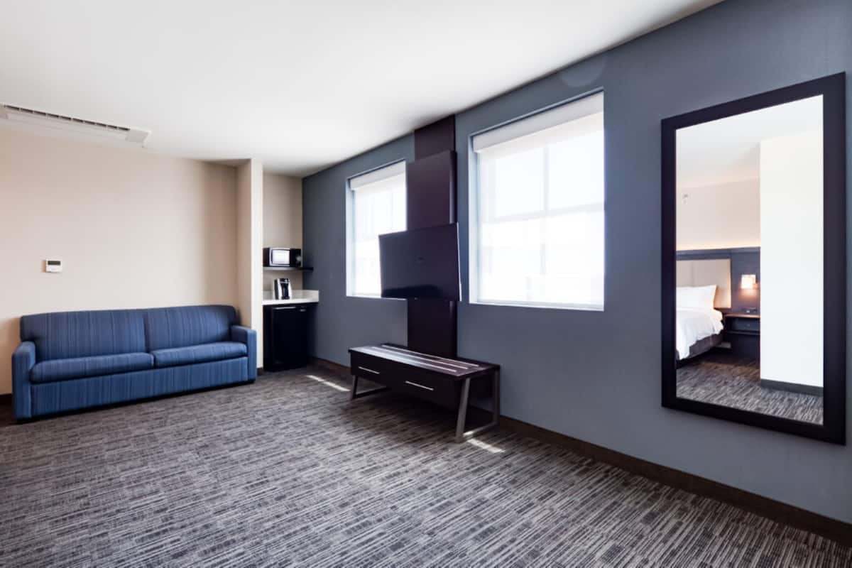 A hotel room with a blue sofa, a small desk, a large mirror, and a view of a bedroom through a window.