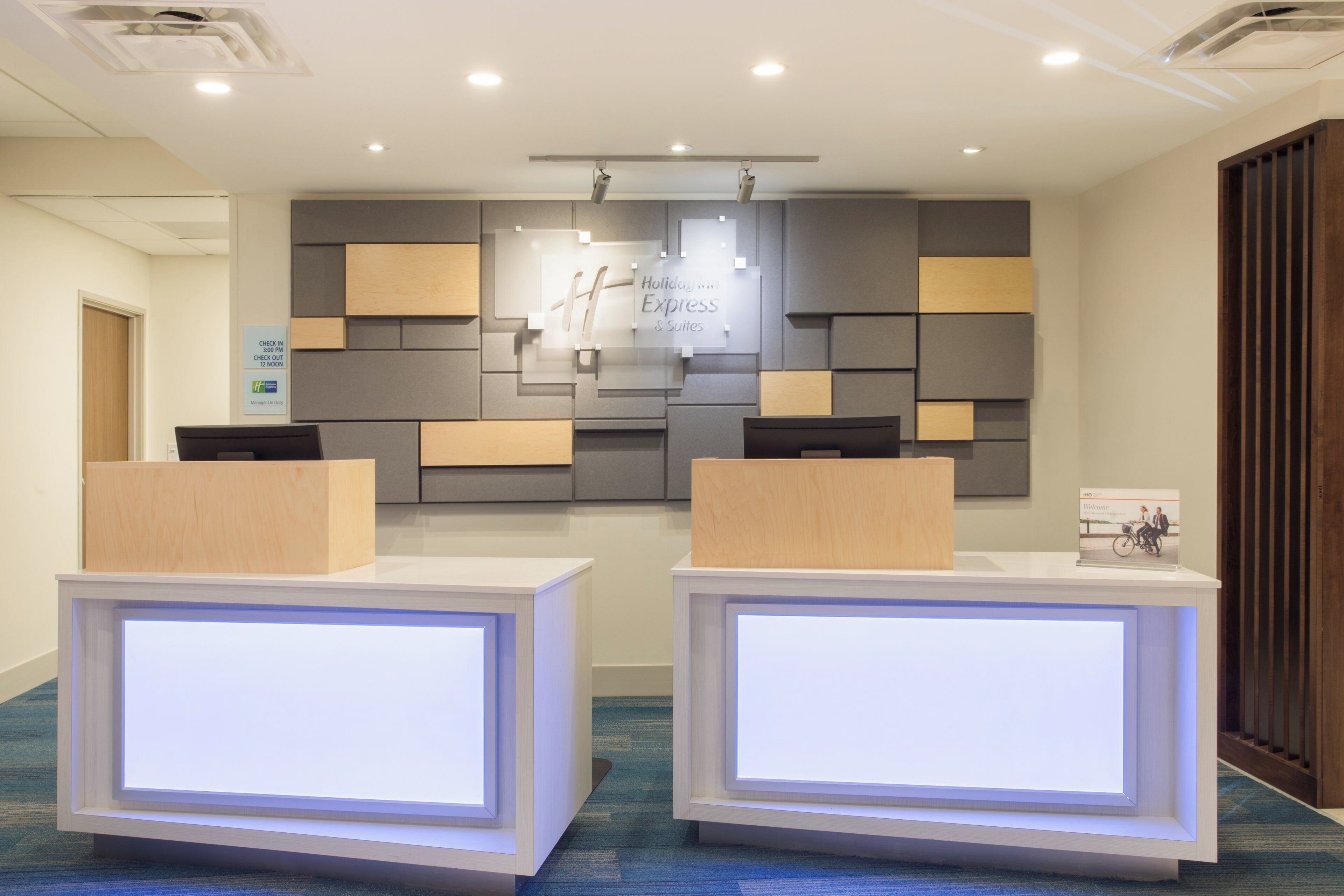 A reception area with a modern design, featuring a check-in counter, a digital display, and a sign indicating the hotel's name and services.
