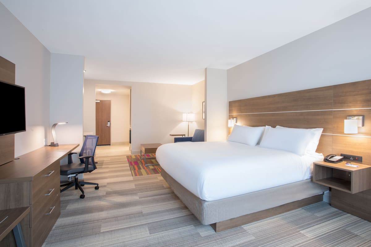 A hotel room with a large bed, a desk with a chair, a TV, and a nightstand with a lamp.