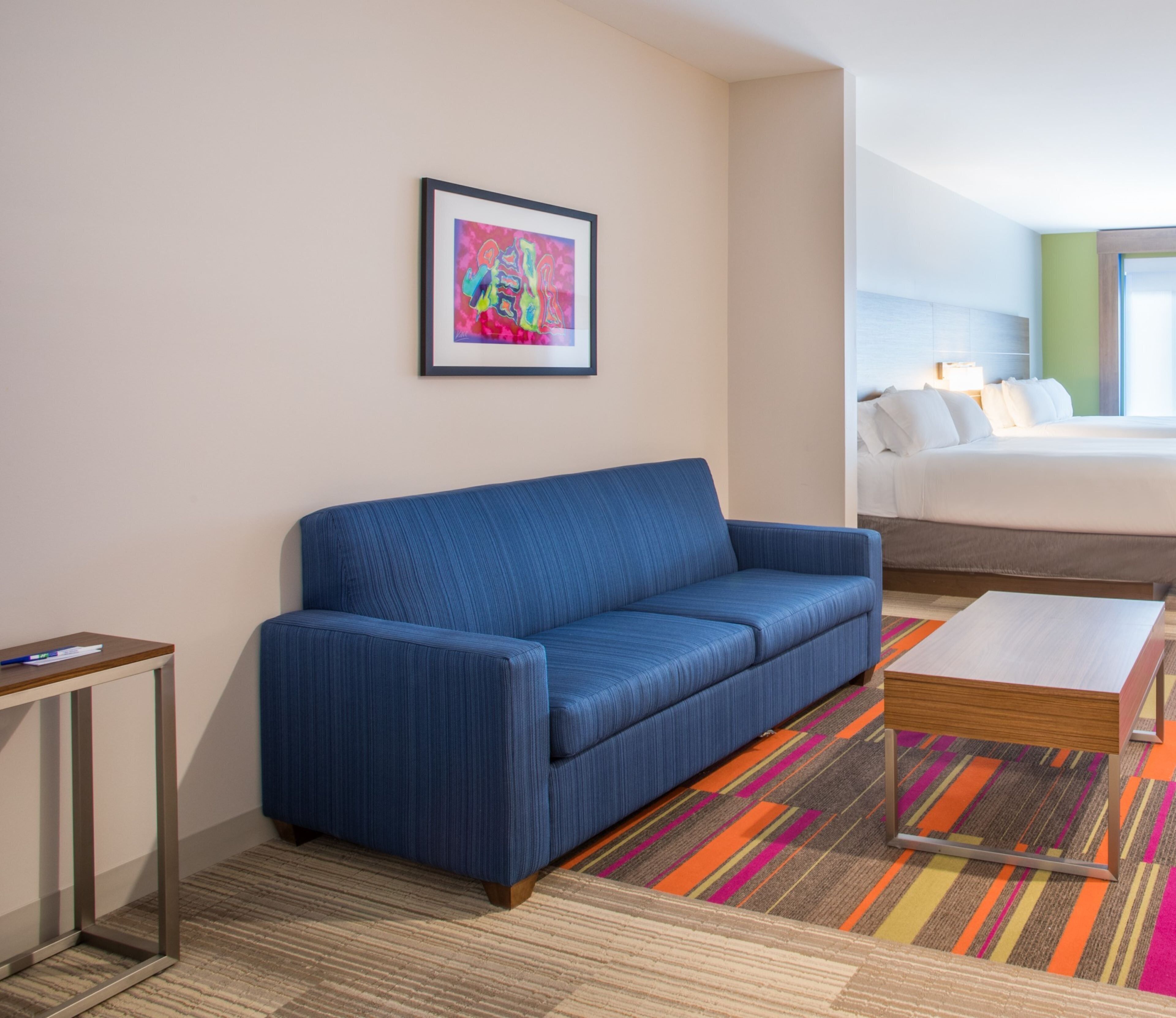Habitación de hotel con un sofá azul, una cama con ropa de cama blanca, un tapete a rayas de colores y un cuadro abstracto enmarcado en la pared.