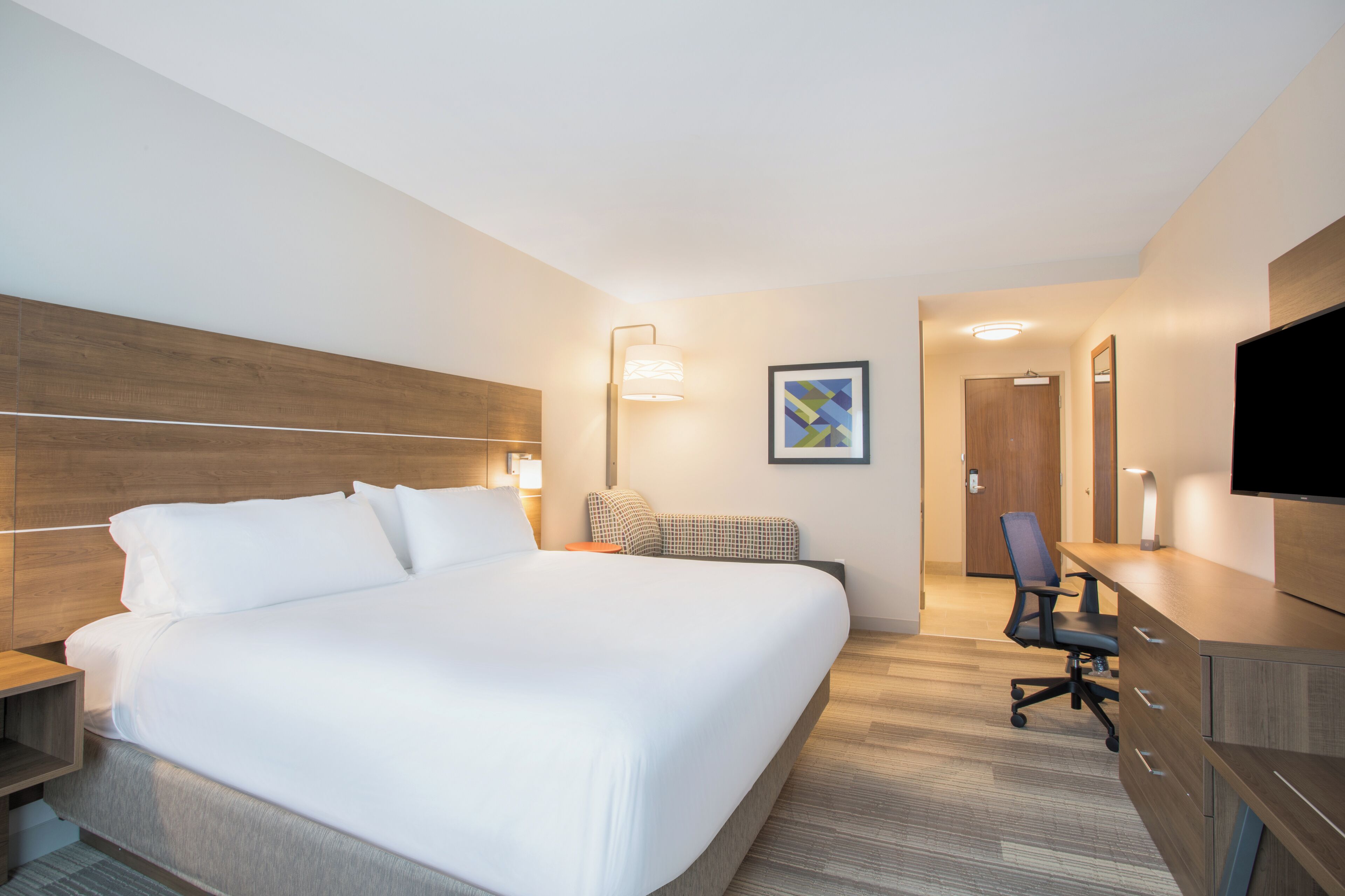 A hotel room with a large bed, a desk, a chair, a television, and a door leading to another room.