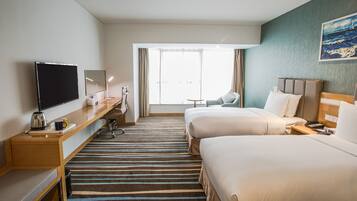 A hotel room with two beds, a desk with a flat-screen TV, a coffee maker, and a large window with curtains.