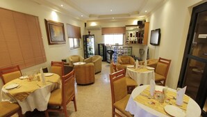 A dining area with round tables set for four, each with white tablecloths and wooden chairs. There are two refrigerators and a bar area with shelves of bottles.