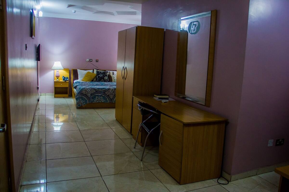 Un dormitorio con cama, escritorio, silla, armario y lámpara de noche.