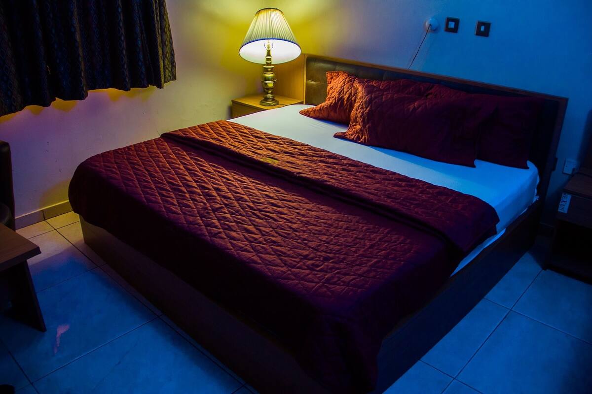 Una cama bien hecha con colcha roja, una lámpara de noche y una mesita de noche de madera.